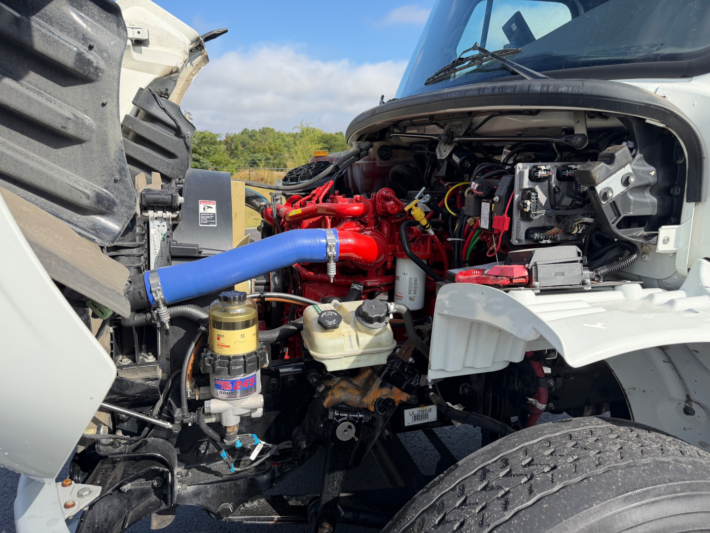 Truck engine bay of a Freightliner M2-106 showing a bright red engine with a blue silicone radiator hose, various hoses, fluid reservoirs, and an exposed alternator area.