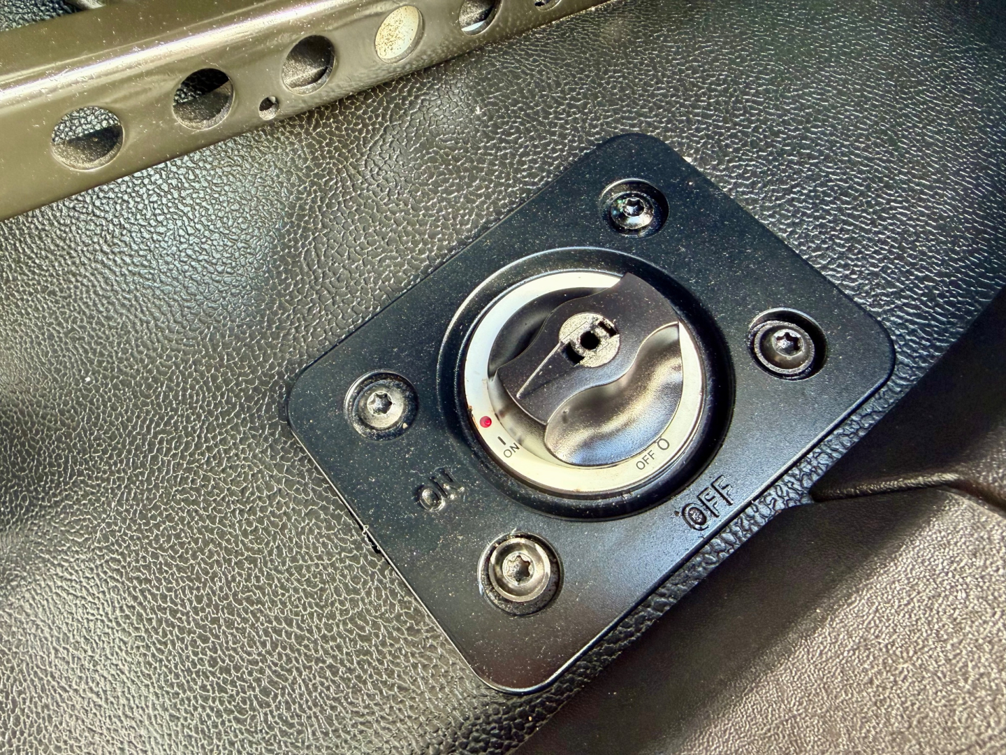 Dashboard view showing a round ignition switch/kill switch on a black panel with four screws and textured surface.