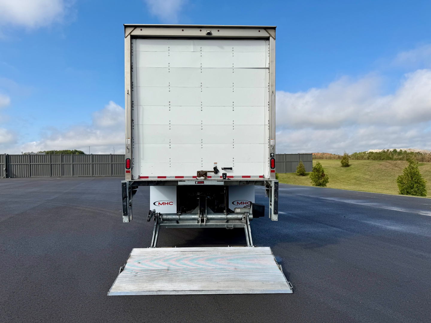 Rear view of a Freightliner M2-106 box truck with ramp deployed. Visible text includes CMBC logo on the bumper and chain hooks. Clear sky and lot in background.