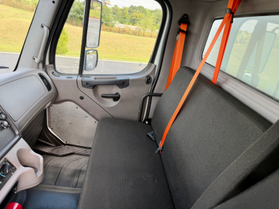 Cab interior of a Freightliner M2-106 showing black bench seat with orange seat belt, grey door panel, and window.