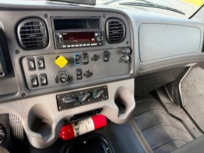 Dashboard cockpit of a Freightliner M2-106 truck showing central audio unit, control switches, climate controls, and a red fire extinguisher mounted under the dashboard.