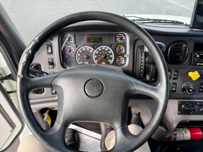 Dashboard and steering wheel of a Freightliner M2-106 cab with instrument cluster showing speedometer, tachometer, and gauges; center console controls visible.