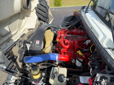 Engine bay of a Freightliner M2-106 showing red engine, blue coolant hose, yellow cap, black air intake, and various hoses and reservoirs.