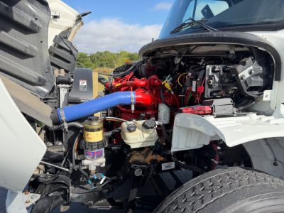 Truck engine bay of a Freightliner M2-106 showing a bright red engine with a blue silicone radiator hose, various hoses, fluid reservoirs, and an exposed alternator area.