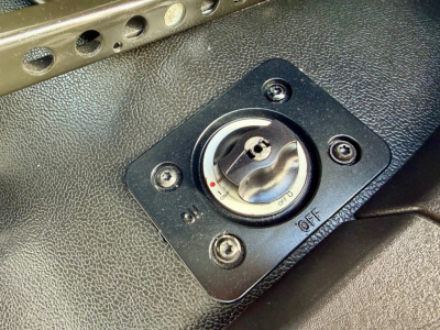 Dashboard view showing a round ignition switch/kill switch on a black panel with four screws and textured surface.