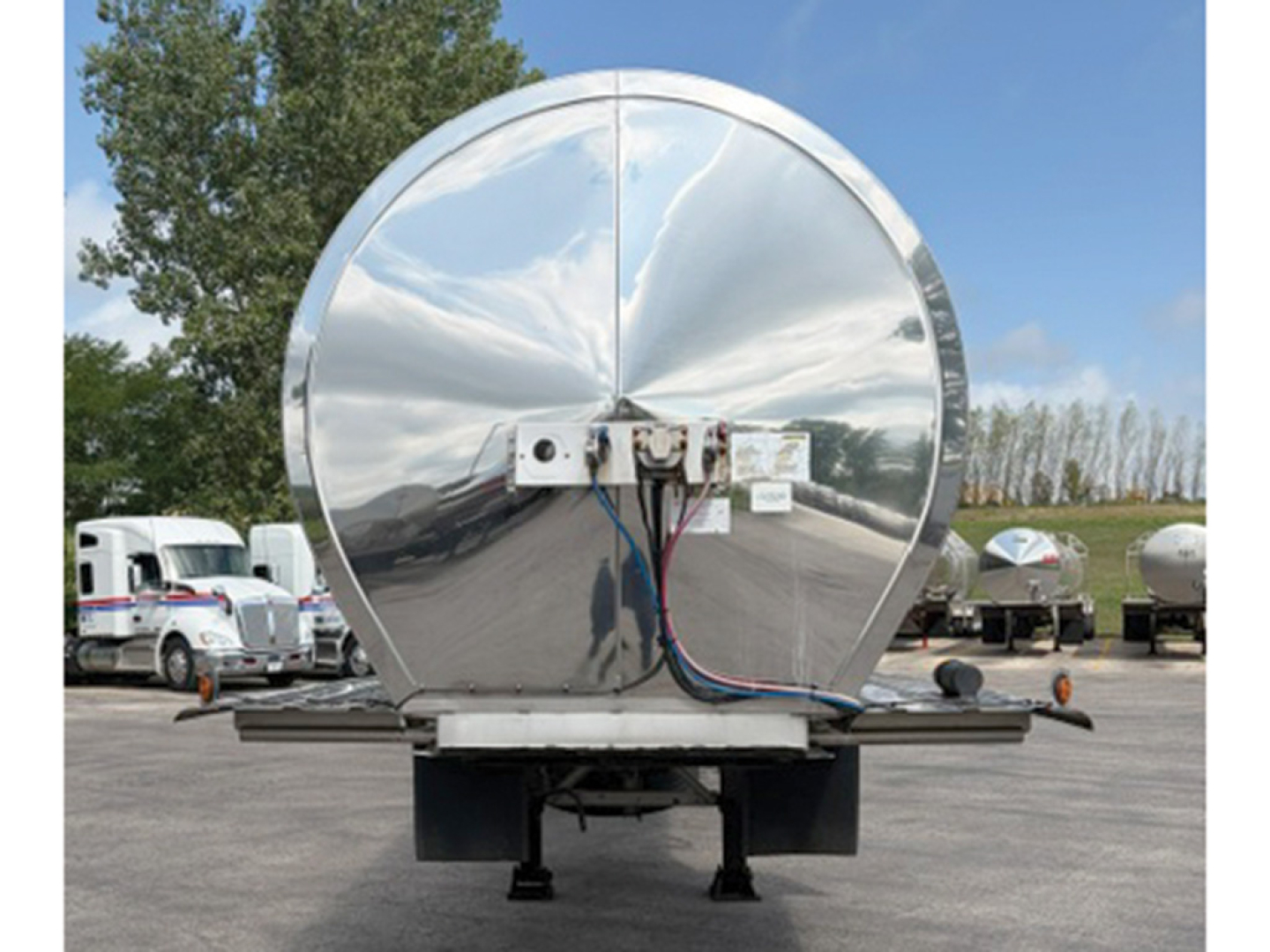Tank trailer rear with polished circular tank and wiring harness mounted on bumper, showing reflection of surrounding lot.
