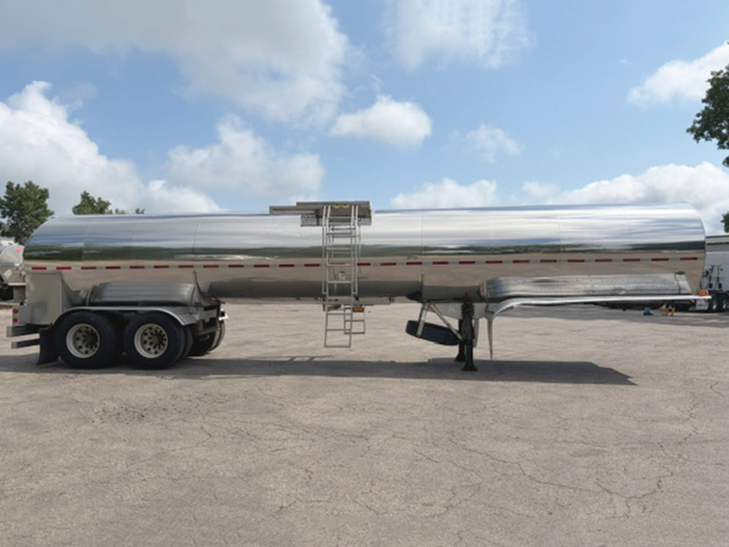 Tank trailer with polished metal cylindrical tank on tandem axle, ladder and access hatch visible, reflection on surface, front hitch and landing gear.
