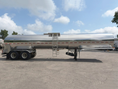 Tank trailer with polished metal cylindrical tank on tandem axle, ladder and access hatch visible, reflection on surface, front hitch and landing gear.