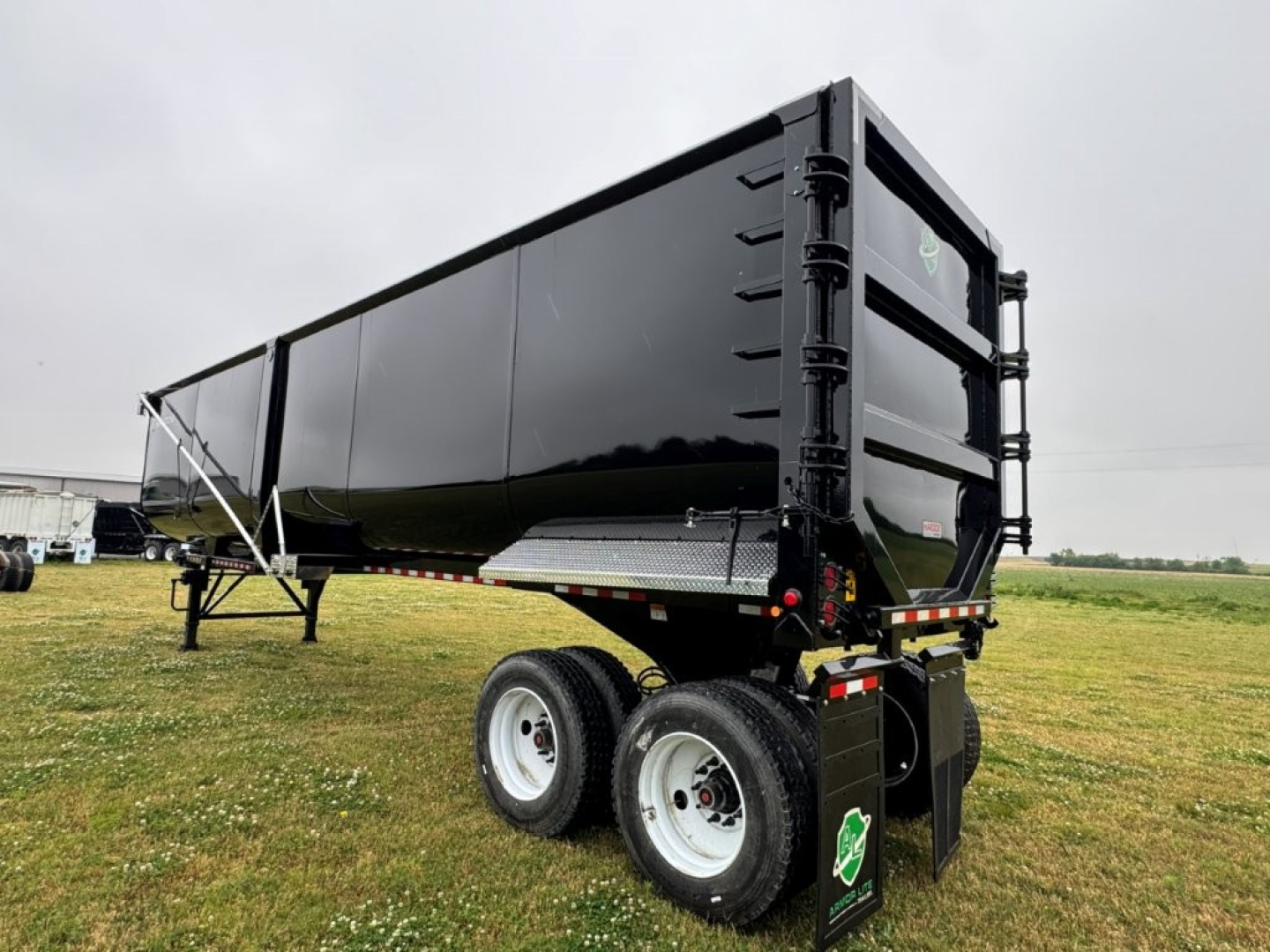Dump trailer with black sides and dual rear axles showing cargo bed and tailgate; green shield sticker on rear; white wheels and support legs.