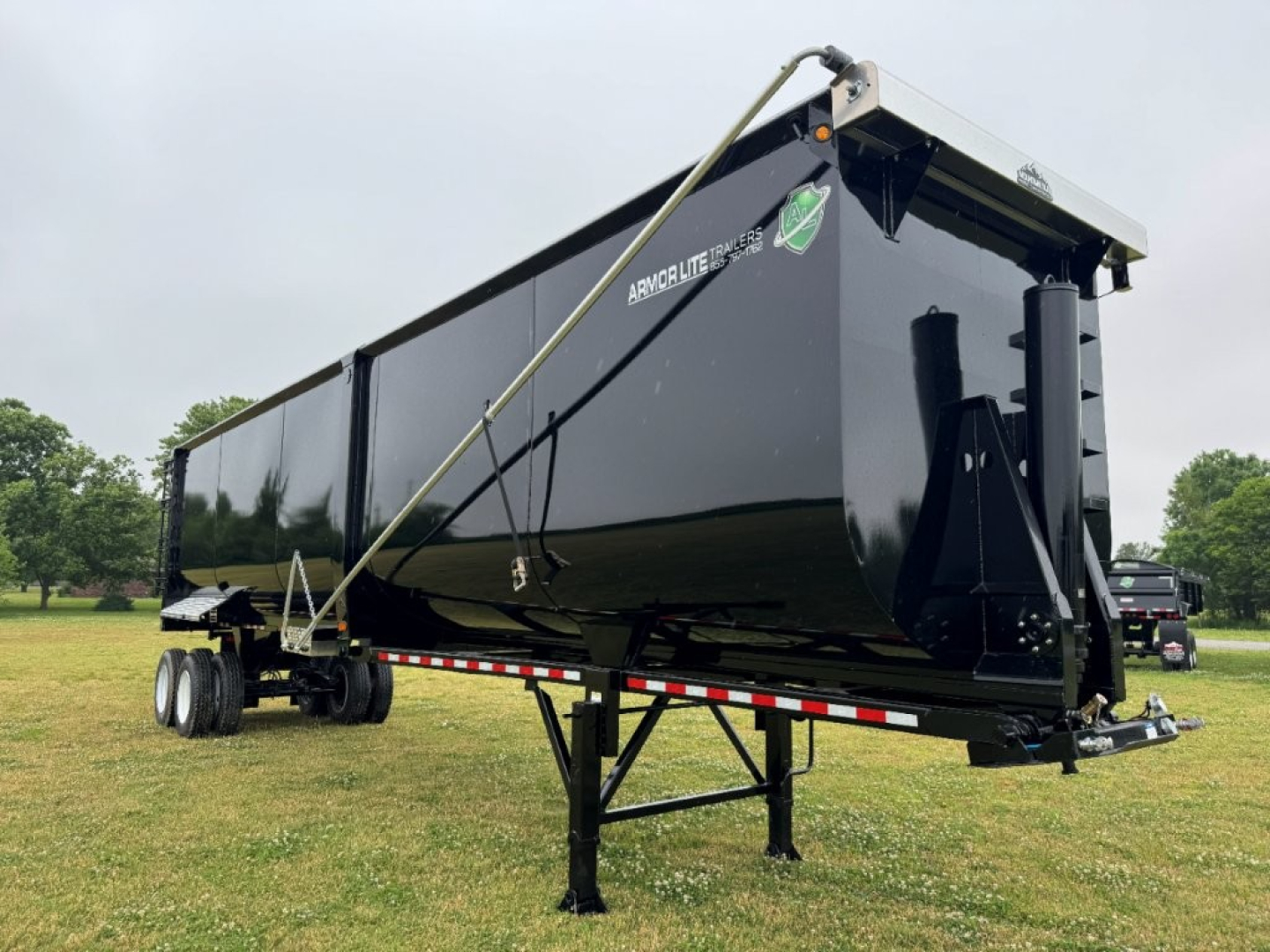 Armor Lite dump trailer showing its black steel body with side rails and a front hitch. Visible text includes ARMORLITE and model details on the side panels and reflective red/white tape along the bottom edge.