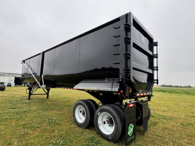 Dump trailer with black sides and dual rear axles showing cargo bed and tailgate; green shield sticker on rear; white wheels and support legs.
