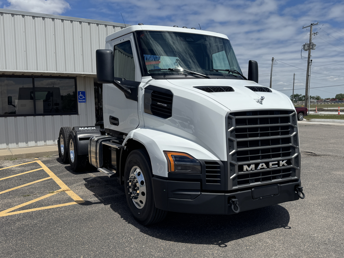 White 2027 Mack AN64T conventional daycab truck front-left view showing grille with MACK emblem, large side mirrors, and vented hood; parking lot, dealership building.