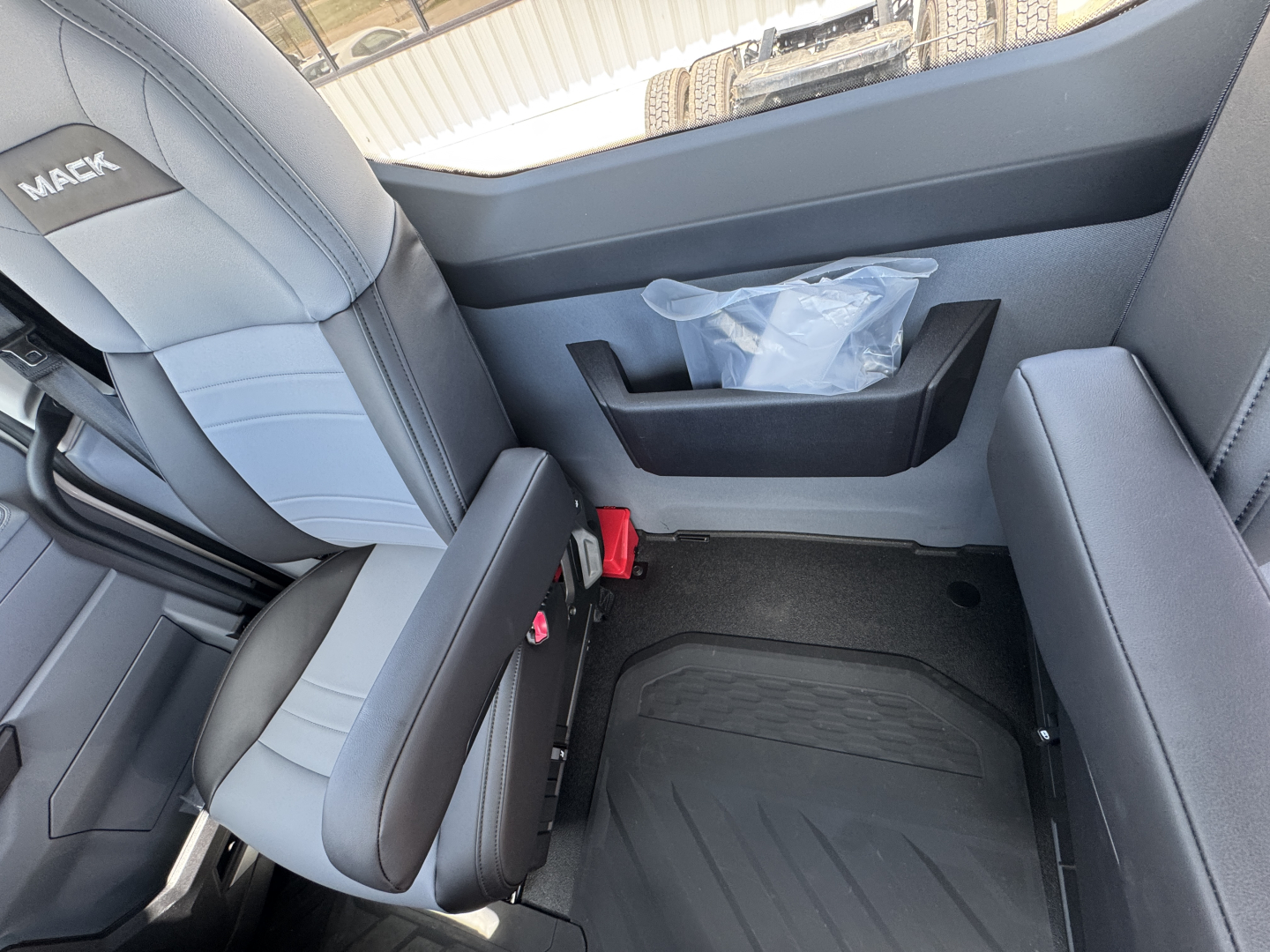 Mack AN64T truck interior showing front passenger seat with gray upholstery, door panel, and storage pocket; part of the cab appears with seat belt buckle and floor mat.