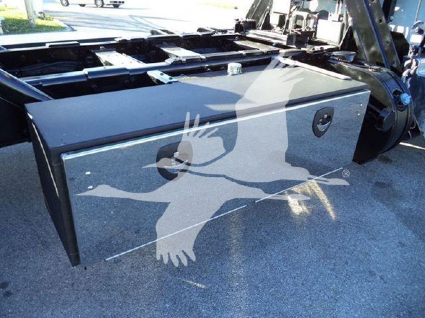 Truck bed toolbox mounted behind cab of a 2020 Peterbilt 337 rollback truck showing a large rectangular aluminum toolbox with a small circular lock, undercarriage frame visible.