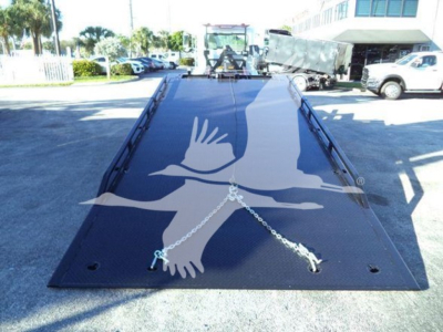 Flatbed tow truck bed with a white gull graphic and chains across, viewed from the front with a Peterbilt rollback in the background.