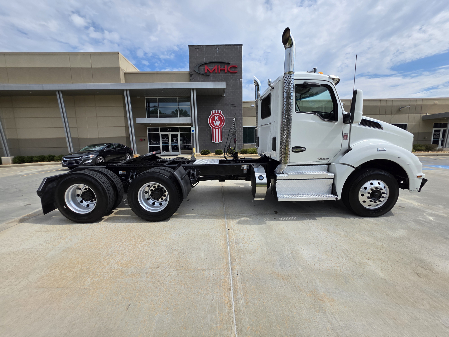 Kenworth T880 tractor cab in side profile view showing chassis with dual rear axles and exhaust stack; white cab, dealership building in background.