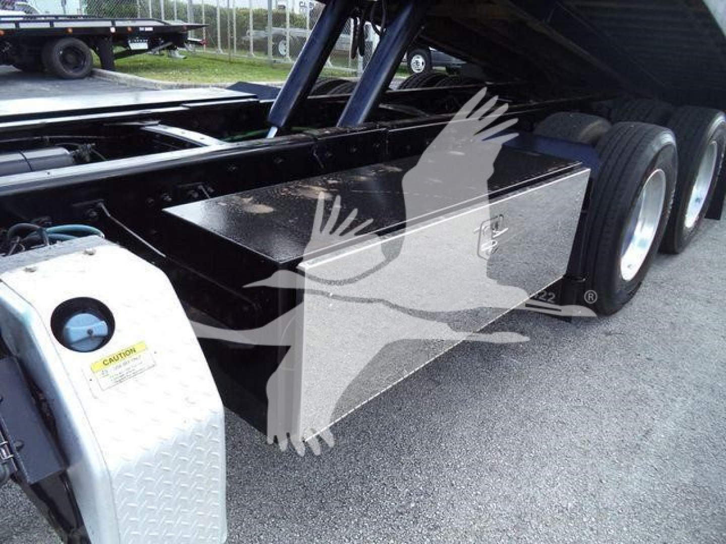 Truck chassis with fuel tank and hydraulic lines visible under the flatbed of a Freightliner M2 106 rollback; warning label on metallic step plate; tires in background.
