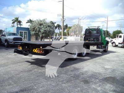 Flatbed rollback tow truck with green cab and long trailer frame, rear view showing dual rear axles and tanker equipment, in a lot.