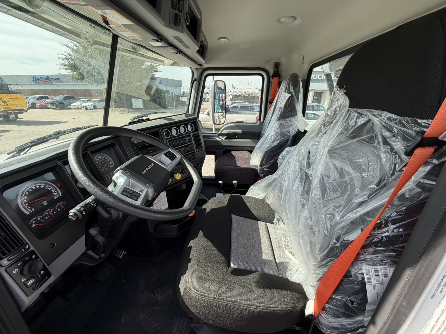 Truck cab interior of new 2026 Mack GR64B showing steering wheel, dashboard with gauges, driver seat wrapped in plastic, orange seat belt.