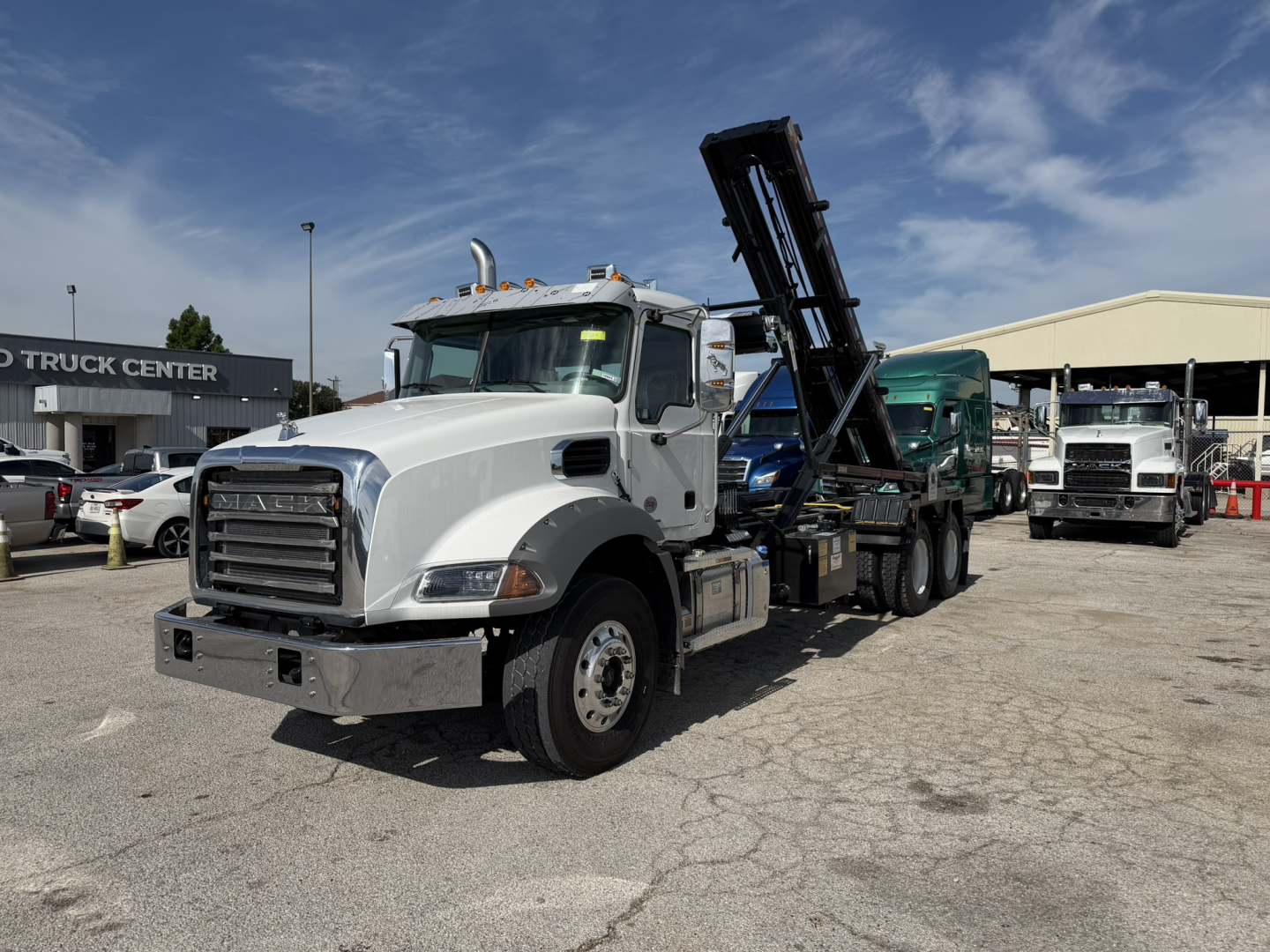 Mack GR64B roll-off truck in white, front-left view showing a raised hydraulic dump/boom, chrome bumper, and wheel arches.