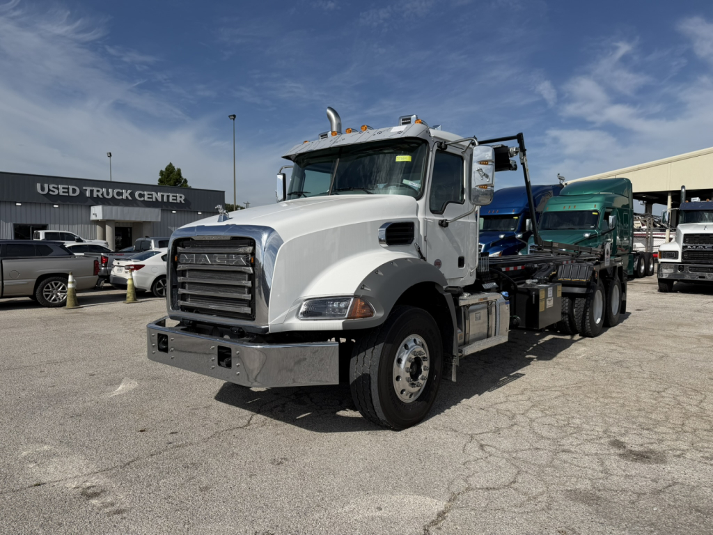 Truck image showing a white Mack GR64B roll-off truck in a dealership lot. Front grille, chrome bumper, dual exhaust stacks, and side mirrors visible.