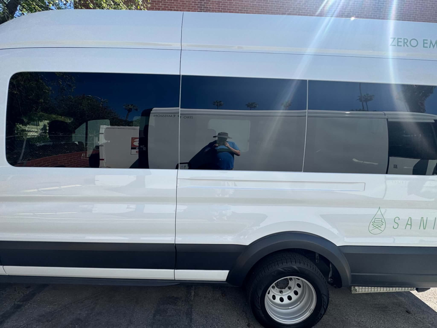 Ford Transit 350 HD van side view showing windows, wheel, and door panels; reflected person taking photo; partial logo and green graphic on rear side.