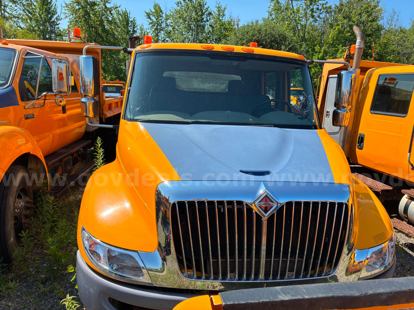 Truck front view of a 2012 International 4300 dump truck, orange cab with chrome grille, other orange trucks parked on either side.