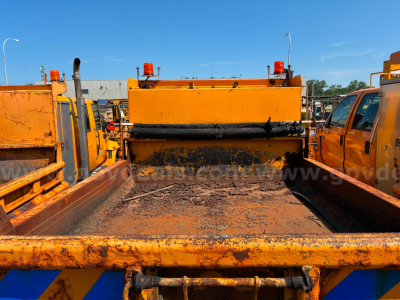 Dump truck bed of an International 4300 chassis showing worn orange bed with rust and loading area, rear-view with yellow equipment, no visible text.