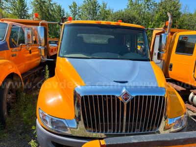 Truck front view of a 2012 International 4300 dump truck, orange cab with chrome grille, other orange trucks parked on either side.