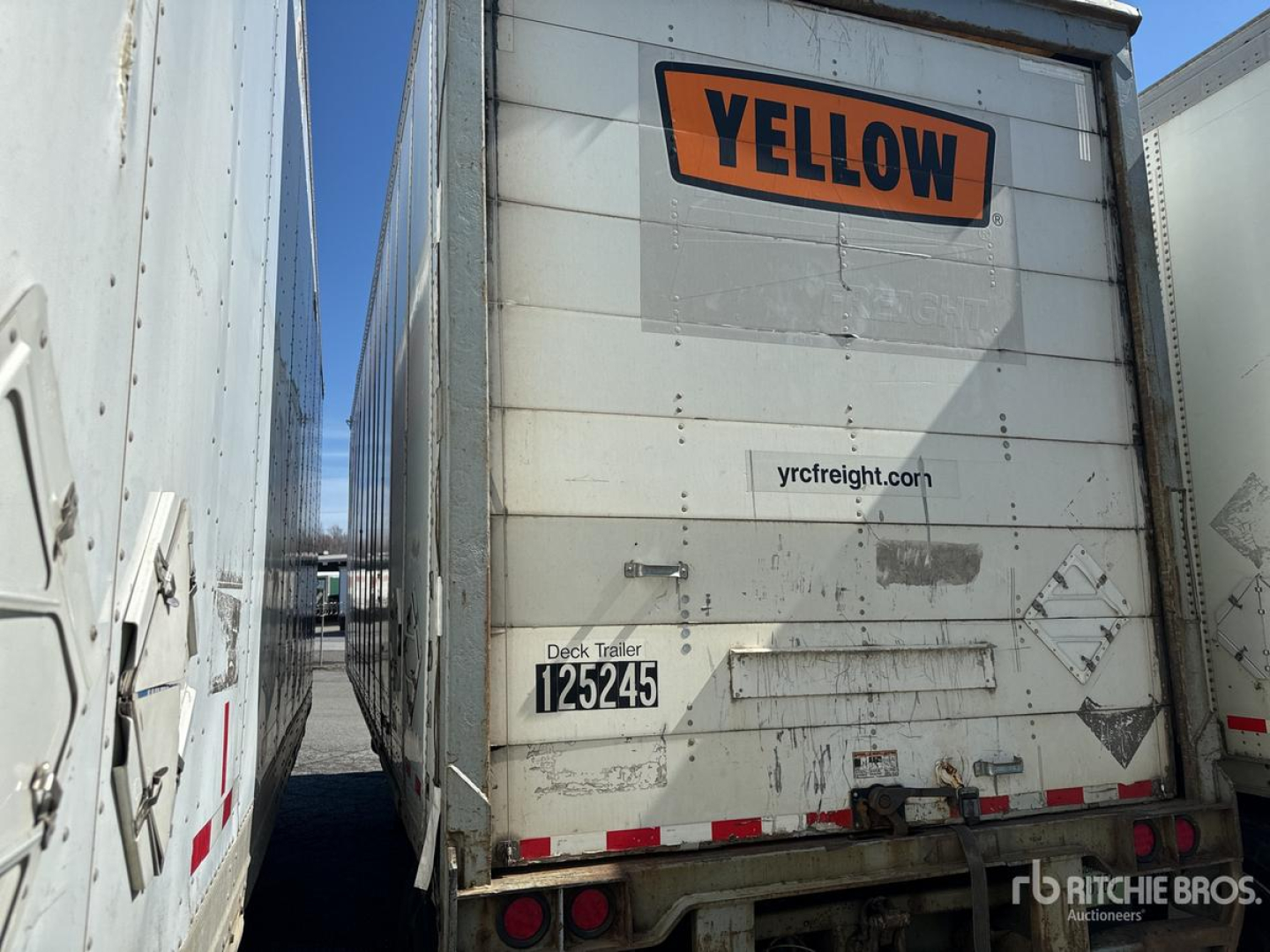 Rear view of a white Wabash trailer with a large orange YELLOW sign at the top and a small website URL on the door, deck trailer label with numbers, and red reflectors along the bumper.