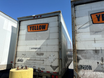 Trailer rear showing YELLOW logo on orange plaque, doors with metal panels, visible numbers and decals, and a yellow bollard in foreground.