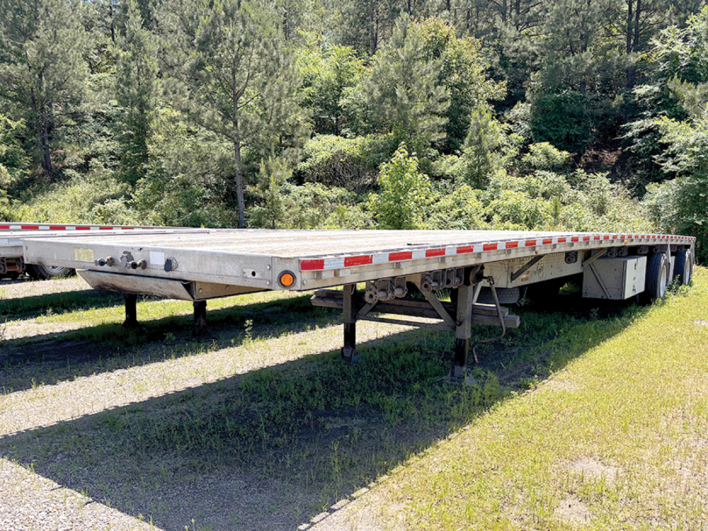 Flatbed trailer (2006 Reitnouer MaxMiser) viewed from side, raised landing gear visible, long silver frame with red/white safety rails on sides.
