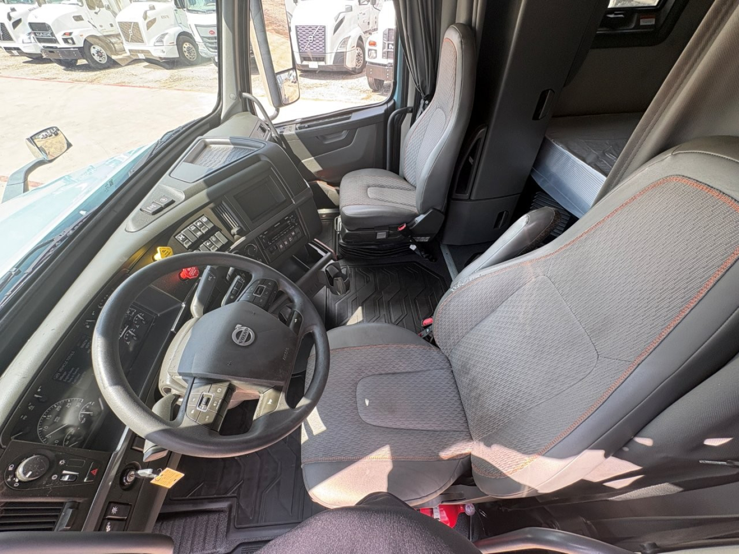 Truck cab interior showing steering wheel, instrument cluster, and driver seat of a Volvo VNL64T760 sleeper with gray seats and red stitching.
