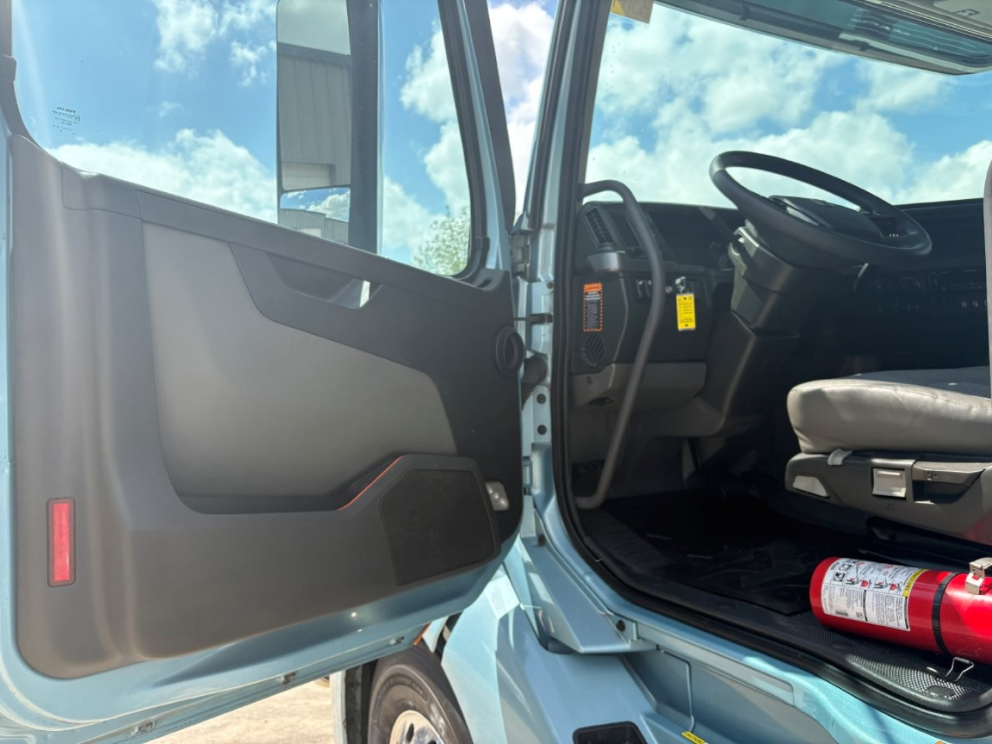 Volvo VNL interior cab doorway view with steering wheel and dashboard visible; passenger seat and floor mat; fire extinguisher stored under seat; open door showing door panel and window frame.