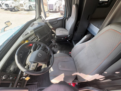 Truck cab interior showing steering wheel, instrument cluster, and driver seat of a Volvo VNL64T760 sleeper with gray seats and red stitching.
