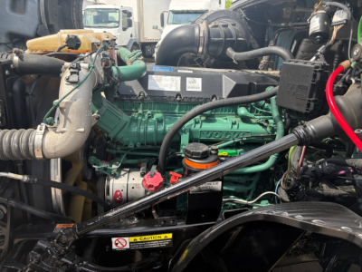 Volvo VNL64T760 engine bay showing green engine block, hoses, belts, and various fluid reservoirs including red cap; label sticker on top; surrounding components and wiring.