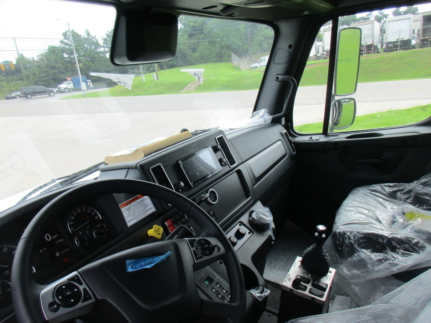Cab interior of a Freightliner M2 106 dump truck showing steering wheel, dashboard, controls, and driver seat wrapped in plastic.