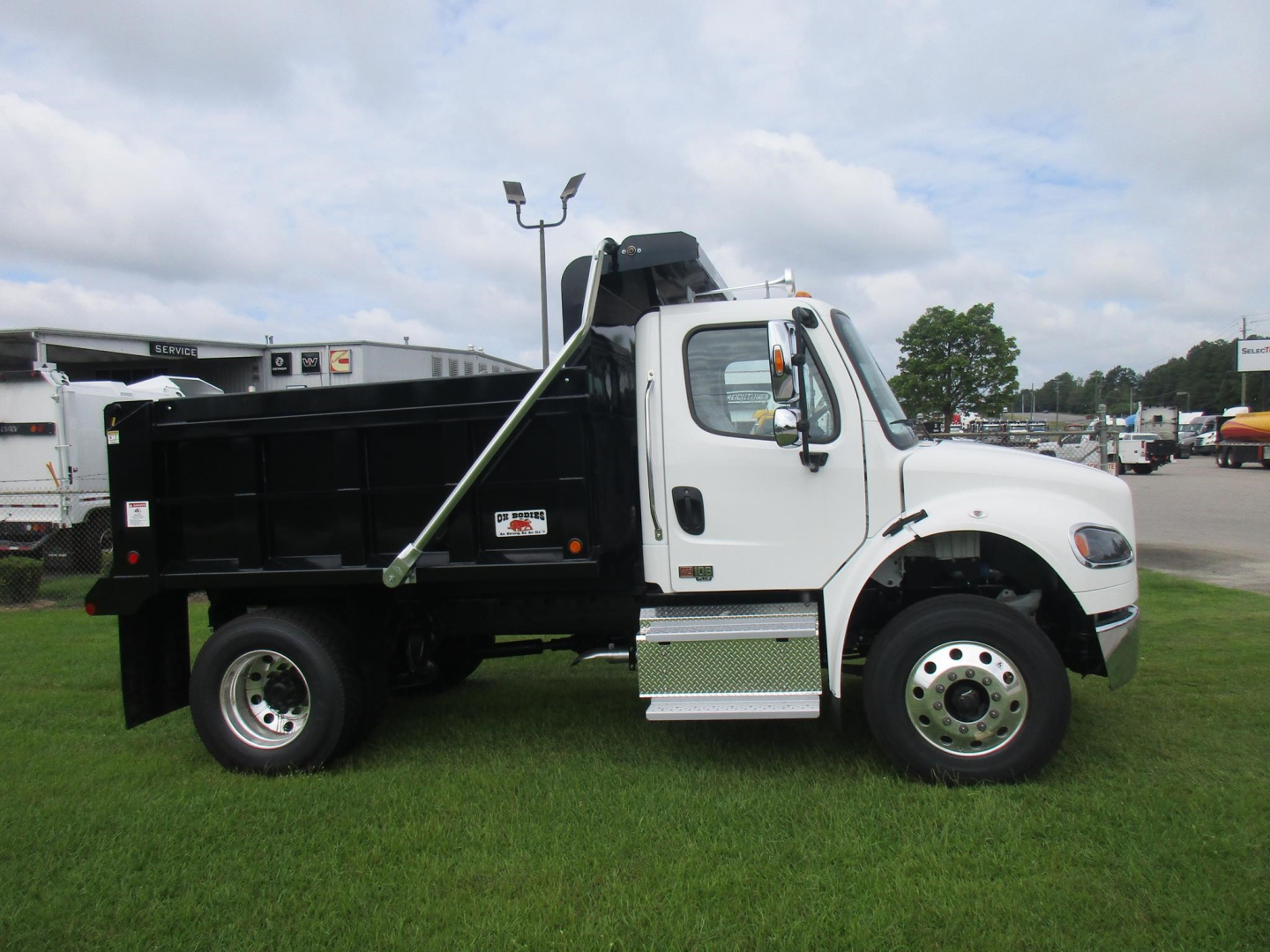 Freightliner M2 106 dump truck side profile showing dumping bed, black container, white cab, chrome steps, and dual rear wheels.
