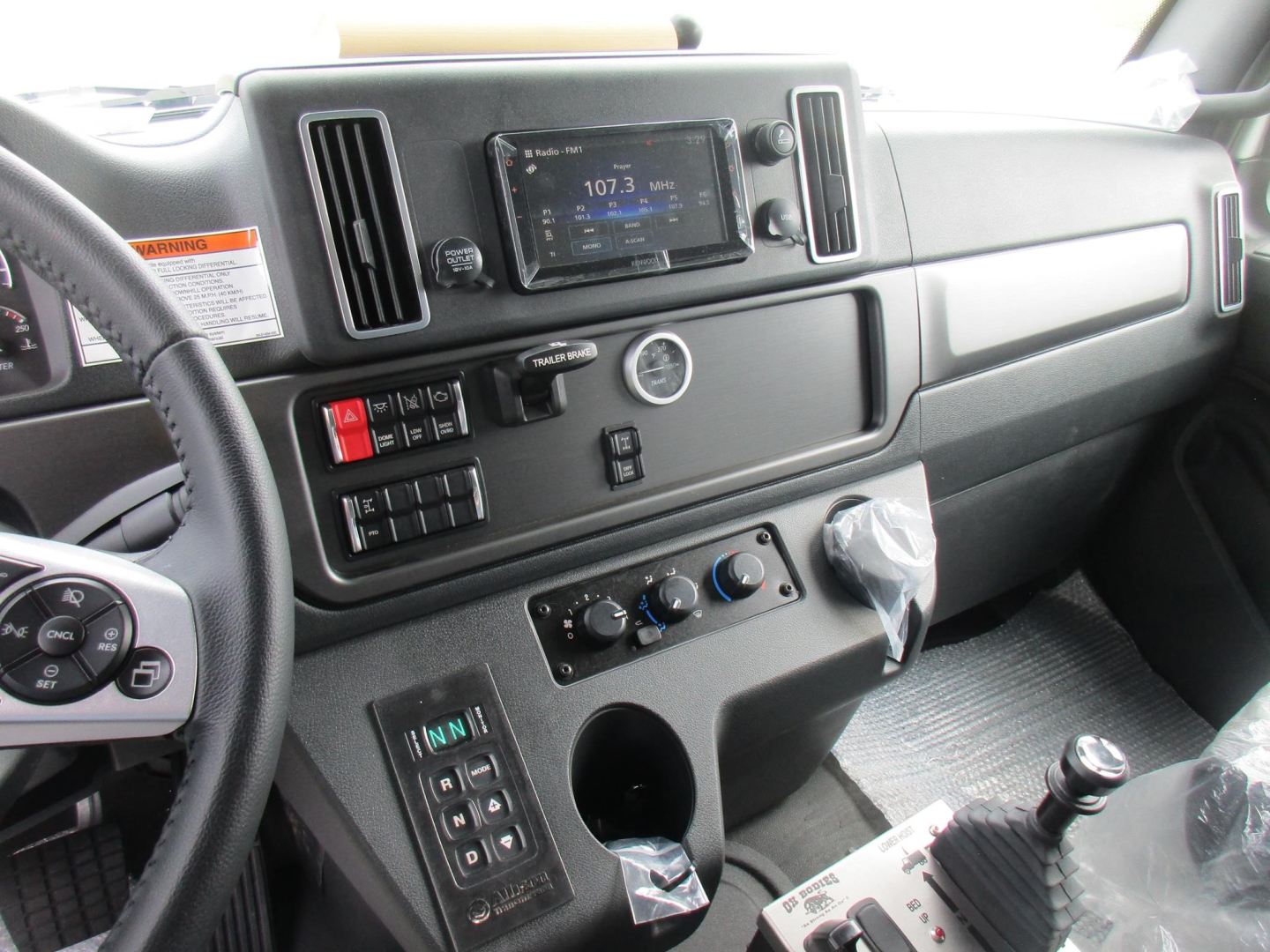 Interior of a Freightliner M2 106 cab showing dashboard with infotainment screen, HVAC controls, gear shifter, and steering wheel.