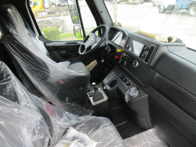 Interior of a Freightliner M2 106 cab showing steering wheel, dashboard, touchscreen monitor, gear shifter, and seats covered in protective plastic.