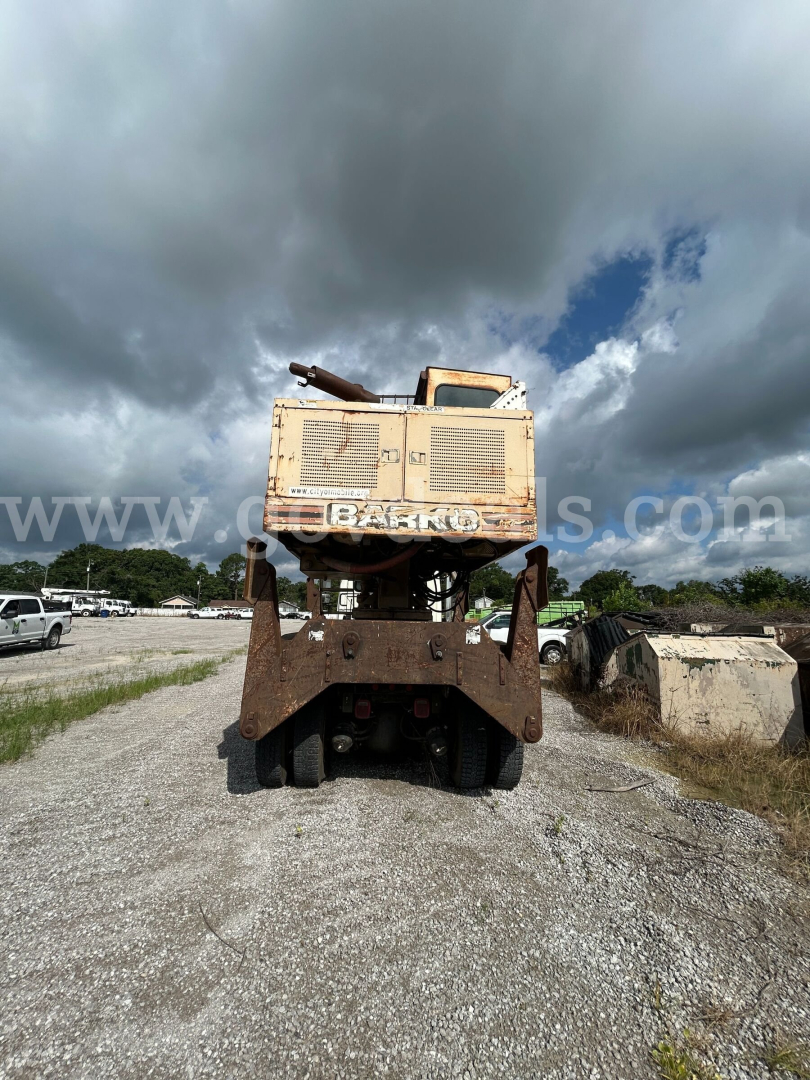 Equipment rear view of a large tracked grapple truck with a rusty undercarriage and beige cab showing the word BAKK? on the back; overcast sky.
