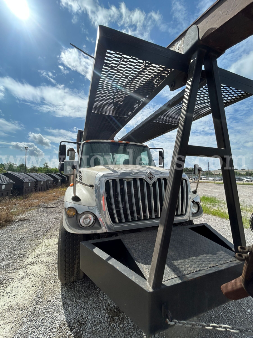 Medium-duty International truck with a large steel grapple attachment mounted above the cab, front grille visible, headlights, and clear blue sky.