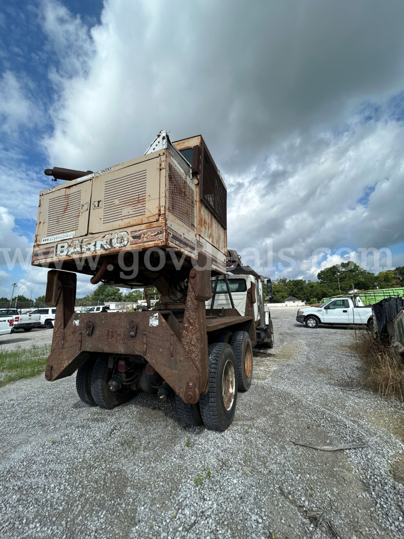 Front rear view of a large grapple truck with a rusted upper equipment carrier lifted over the cab; visible tires; some text on the truck body including a watermark across the window.