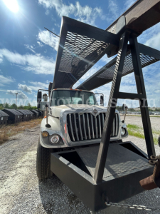 Medium-duty International truck with a large steel grapple attachment mounted above the cab, front grille visible, headlights, and clear blue sky.