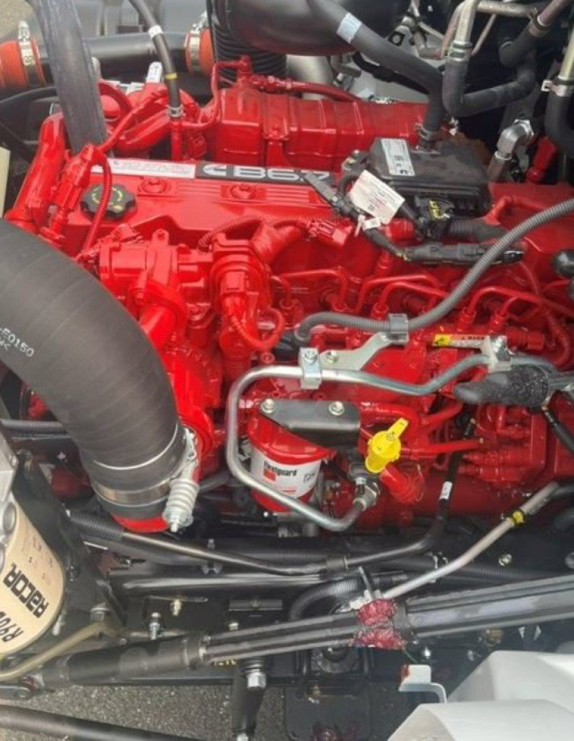 Red engine bay of a 2027 Hino L6 cab chassis showing engine components, hoses, and a yellow oil dipstick; visible labels and fittings on the red motor.