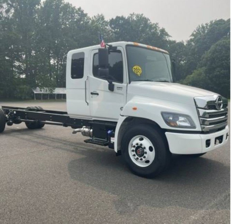White 2027 Hino L6 cab chassis truck in showroom lot showing front and left side with chassis exposed.