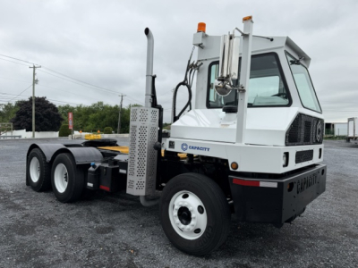 Capacity TJ6500 truck showing side view with cab and chassis, white with black accents, orange beacon lights, exhaust stack, and large wheels.