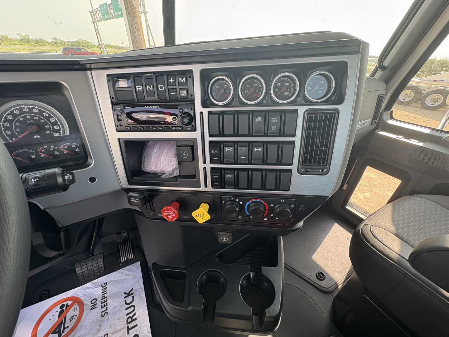 Truck cab interior showing instrument cluster, dashboard controls, and gear shifter in Mack GR64FT. Visible text on controls includes R N D P and other switch labels.