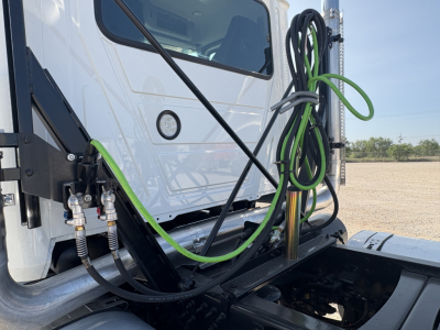 Truck cab exterior showing green and black hoses connected near front, date cab area, Mack GR64FT chassis, daytime outdoor setting.