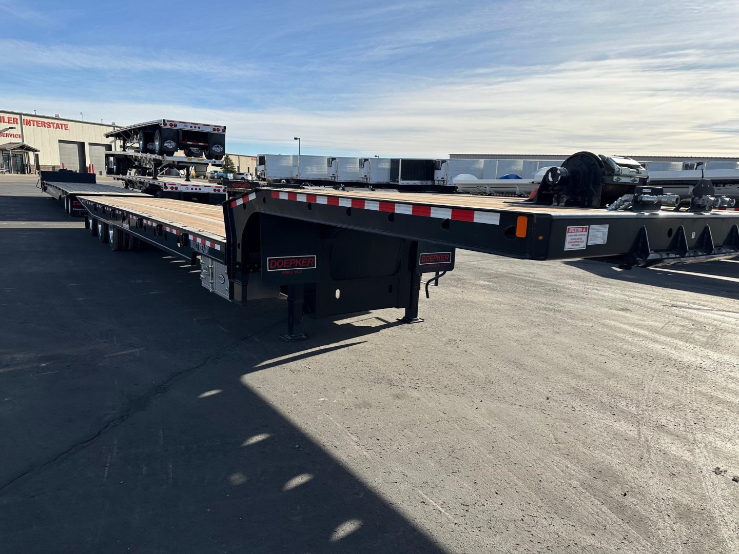 Doepker RT trailer in an outdoor lot, showing a long flatbed trailer with landing gear and reflectors; visible brand stickers say Doepker and a stacked equipment in background.